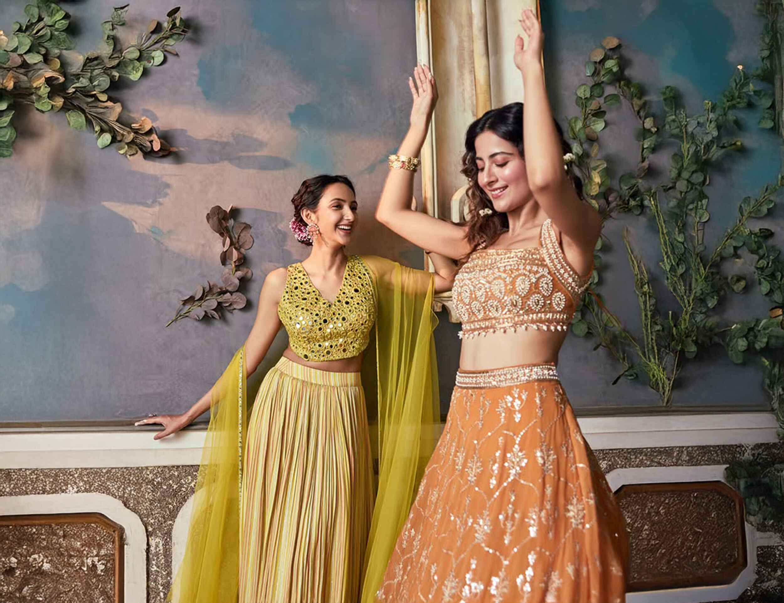 Women in vibrant festive lehengas &ndash; one in a yellow mirror-work outfit and the other in an orange embroidered lehenga &ndash; dancing joyfully for Ganesh Chaturthi 2025 celebrations.